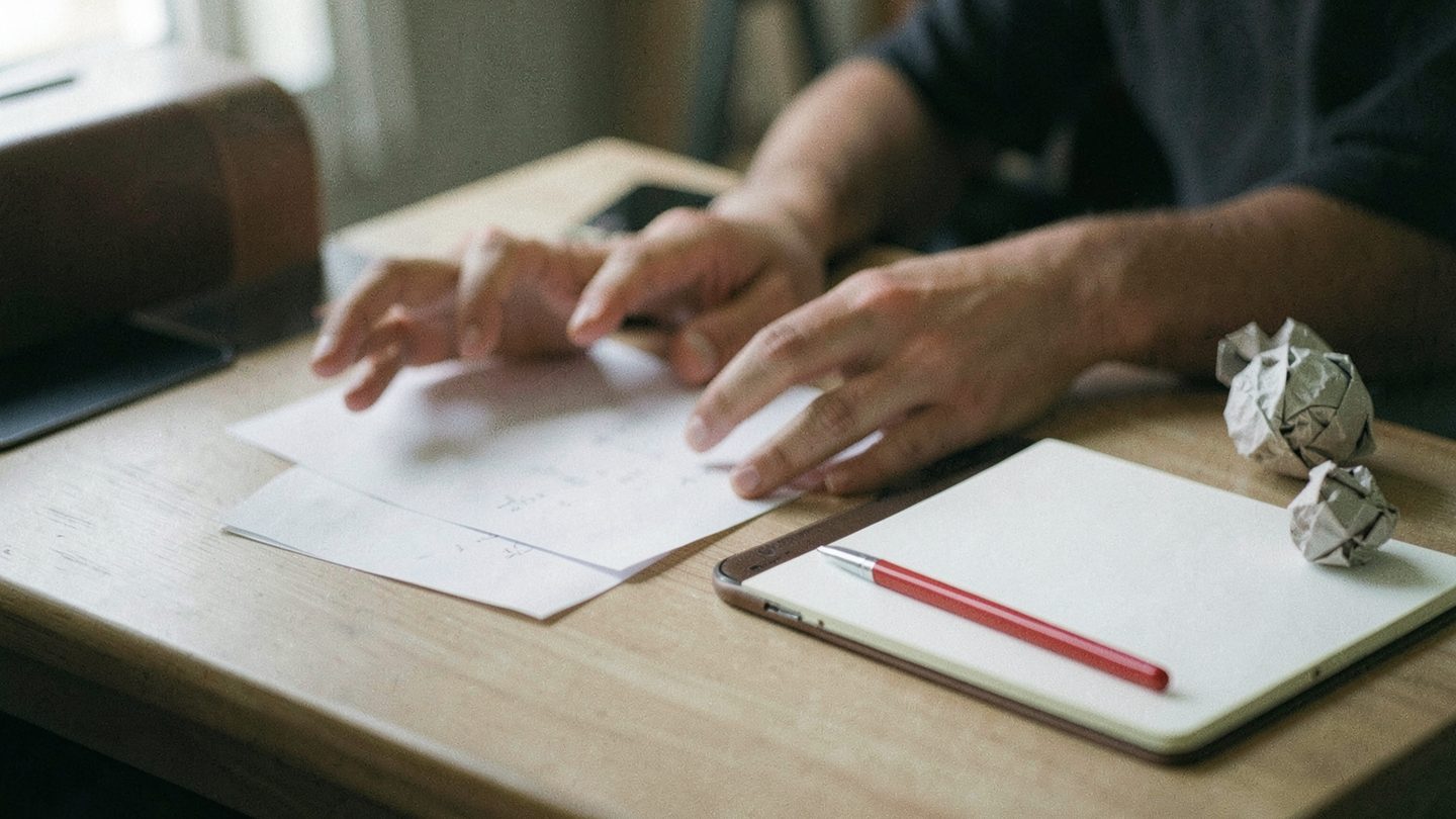 A realistic editorial-style desk scene in landscape orientation showing hands over blank papers, a face-down phone, a red pen, a clean open notebook, and crumpled paper balls in soft window light.
