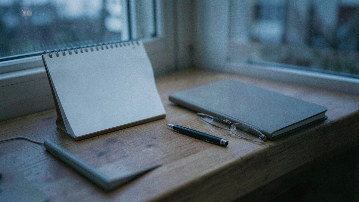 A blank desk calendar pad and closed notebook on a wooden table beside a window at dusk, with a pen and reading glasses nearby.