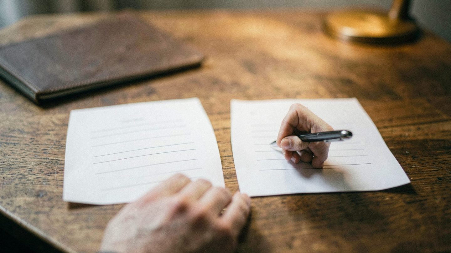 A close-up editorial photo of a hand hesitating above two blank paper lists on a desk, with a softly blurred lamp and notebook in the background, suggesting indecision.