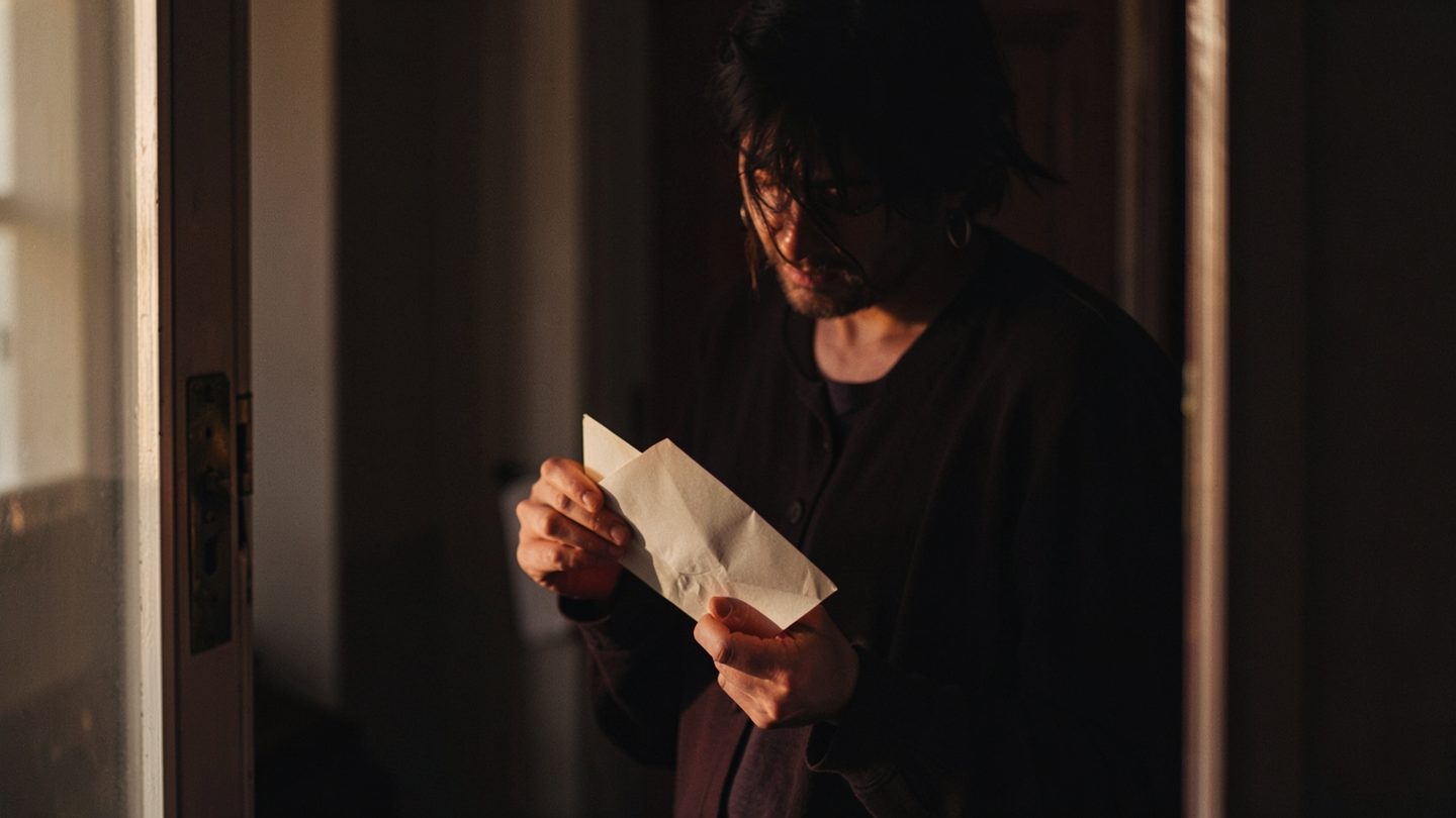 A person in a doorway holding a sealed envelope, evoking privacy and slow disclosure
