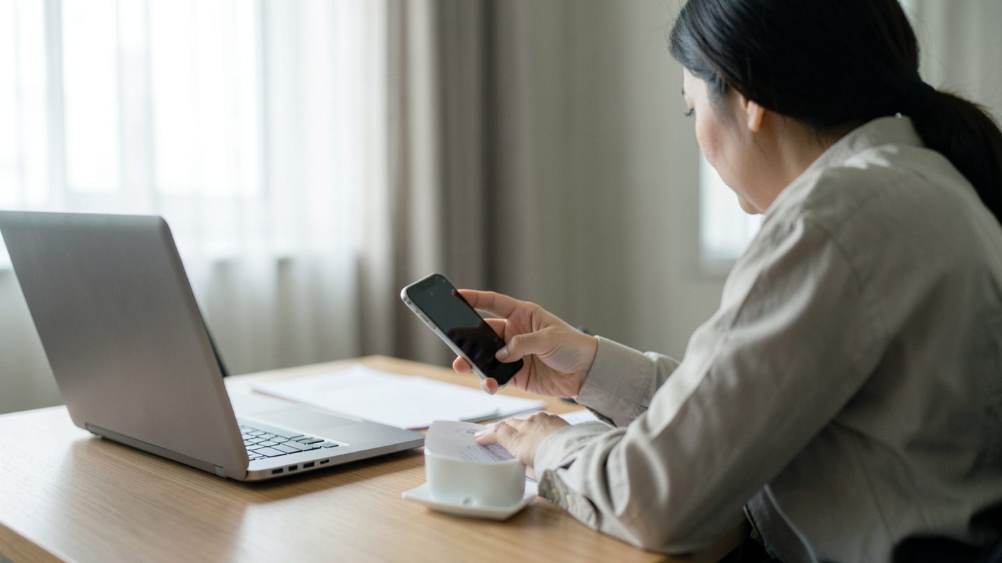 A person sits at a desk in a quiet office, holding a smartphone and hesitating before sending a message, with a closed laptop, blank papers, and a coffee cup nearby.