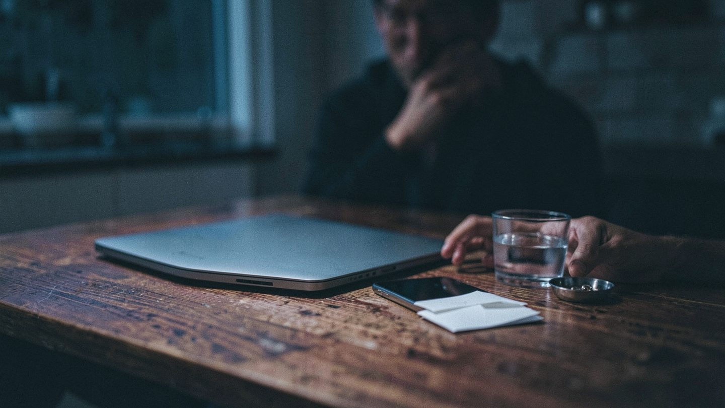 A moody nighttime kitchen table scene with a face-down phone, unopened envelopes, a glass of water, and a ring dish while a thoughtful person sits in partial shadow nearby.